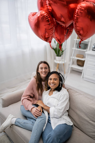 mujeres lesbianas positivas y multiétnicas sentadas en el sofá cerca de globos en forma de corazón en el día de San Valentín  - Foto, Imagen