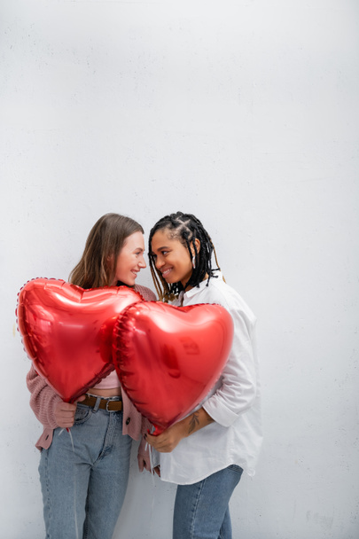 Sevgililer gününde kırmızı balon tutan pozitif ve etnik çeşitlilikte lezbiyen kadınlar gri renkte izole edilmiş.  - Fotoğraf, Görsel
