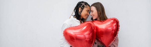 femmes lesbiennes positives et multiethniques tenant des ballons rouges en forme de cœur isolés sur une bannière grise - Photo, image