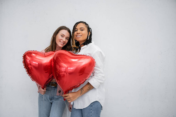 vidám és fajok közötti leszbikus nők, akik szív alakú lufikat tartanak Valentin-napon elszigetelve a szürkén  - Fotó, kép