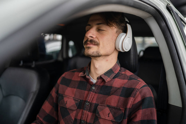 One man young adult caucasian male sit in the car on the seat with headphones listen guided meditation relaxation practicing mindfulness or listen music or podcast real people copy space eyes closed - Foto, immagini