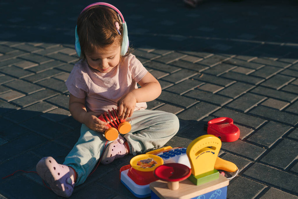 Dziewczynka siedzi w ogrodzie o zachodzie słońca bawiąc się wieloma zabawkami, które rozwijają umiejętności. Naturalne lato. Zabawki edukacyjne. Szczęśliwa rodzina. - Zdjęcie, obraz
