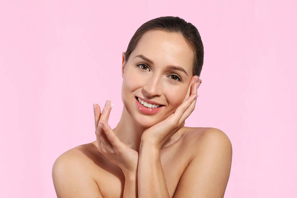 Studio portrait of young beautiful woman with long black hair tied in ponytail applying anti aging cream to her face. Close up, copy space, isolated pink background. - Photo, image