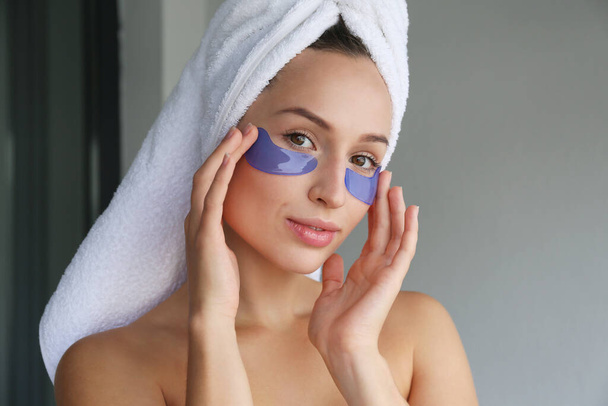 Young woman wearing a towel after showering, pampering the under eye patch for dark circles treatment. Female applying cosmetic products to decrease puffiness under her eyes. Close up, background. - Фото, зображення