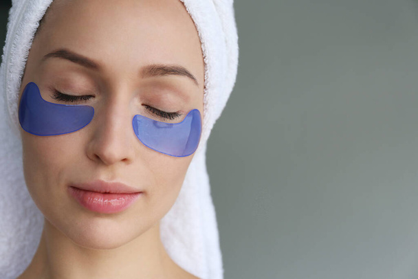 Young woman wearing a towel after showering, wearing eye patch for dark circles treatment. Female applying cosmetic products to decrease puffiness under her eyes. Close up, background. - Photo, Image
