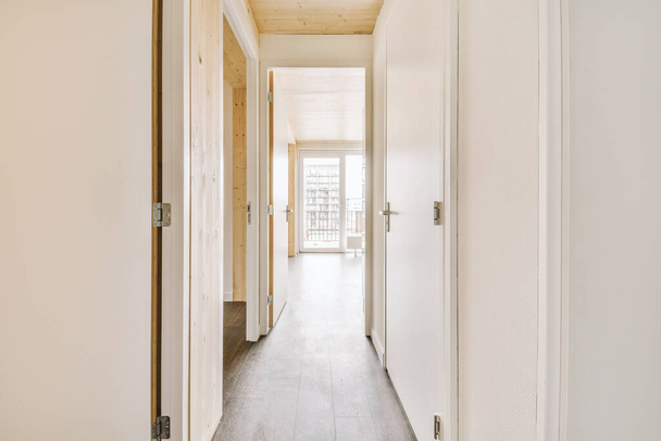 Pasillo estrecho de luz con muchas puertas en paredes blancas y lámpara brillante sobre suelo de madera - Foto, Imagen