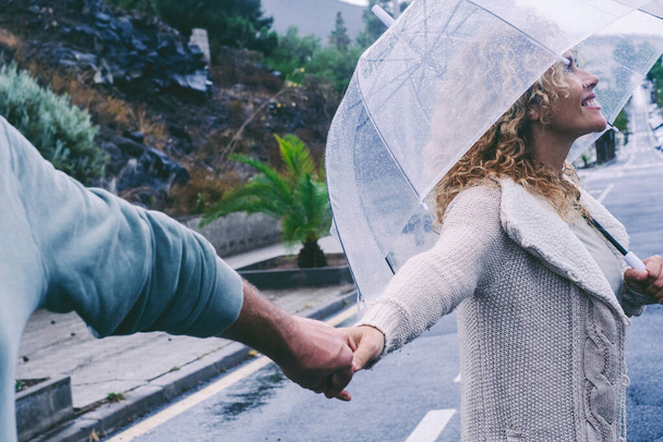 Gelukkig volwassen stel genieten van het leven in een regenachtige dag samen in romantische vrijetijdsbesteding buitenactiviteiten onder een paraplu met straat weg op de achtergrond. Man en vrouw hand in hand onder de regen - Foto, afbeelding