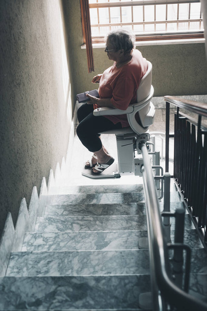 staruszka z problemami z nogami używa krzesła elektrycznego do wspinania się po schodach w domu. Technologia wspomagająca starsze życie dla ludzi - Zdjęcie, obraz