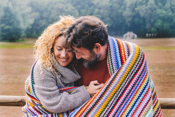 Romantic adult couple in love hug with tenderness under a colorful wool cover in outdoor park. Concept of winter leisure activity for man and woman in relationship. People embracing and enjoying - Photo, Image