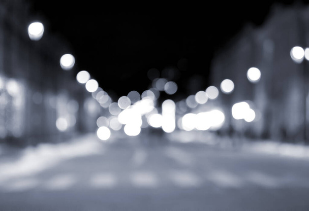 Fondo borroso. Ciudad nocturna. Silueta borrosa de edificios, manchas bokeh de linternas brillantes, siluetas borrosas de personas caminando por la calle por la noche. Plaza de la ciudad, cielo nocturno negro Color azul - Foto, Imagen