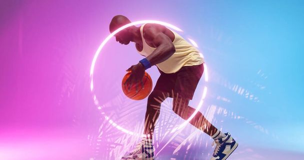 Side view of bald african american player dribbling basketball by illuminated circle and plants. Copy space, composite, sport, competition, illustration, glowing, nature, shape and abstract concept. - 写真・画像