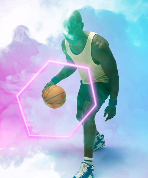 African american male player dribbling basketball by illuminated hexagon on blue smoky background. Copy space, composite, sport, competition, illustration, glowing, shape and abstract concept. - 写真・画像