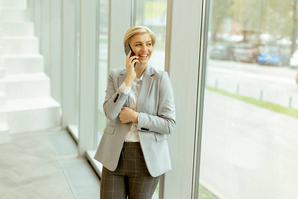 Femme d'affaires utilisant un téléphone portable tout en se tenant sur le couloir de bureau moderne - Photo, image