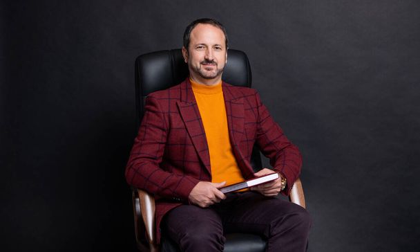 cheerful businessman sitting in office chair with planner isolated on black background. businessman sitting with planner in studio. sitting businessman holding planner. photo of businessman sitting - Photo, image