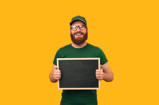 Счастливый бородатый мужчина в форме курьера и в очках держит доску на жёлтом фоне. - Фото, изображение