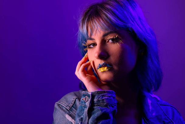 Portrait of young stylish woman posing indenim jacket, blue and yellow tones of clothes in Ukrainian colors style, flowers art on lips - 写真・画像