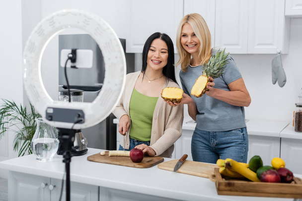Χαρούμενοι διαφυλετικοί φίλοι που κόβουν φρούτα κοντά στο μπλέντερ και το smartphone στην κουζίνα  - Φωτογραφία, εικόνα