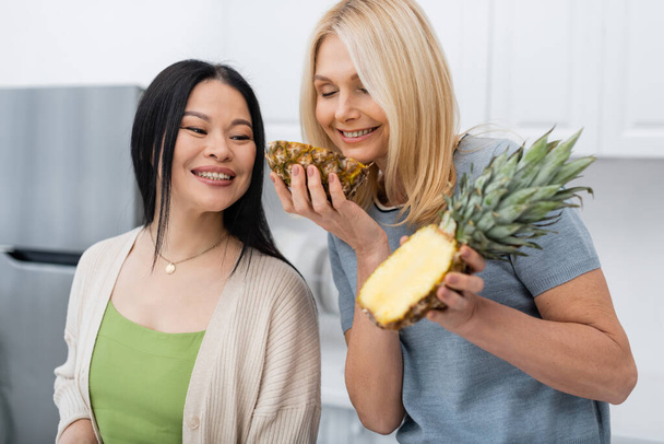 Uśmiechnięta kobieta pachnąca świeżym ananasem w pobliżu azjatyckiego przyjaciela w kuchni  - Zdjęcie, obraz