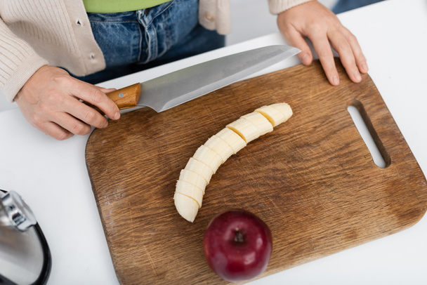 Vista dall'alto della donna che tiene il coltello vicino a banana e mela sul tagliere  - Foto, immagini