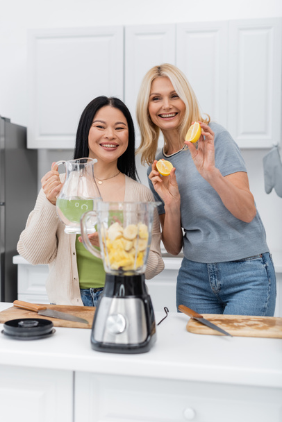 Positive interracial friends holding fruits and water near blender in kitchen  - Photo, Image