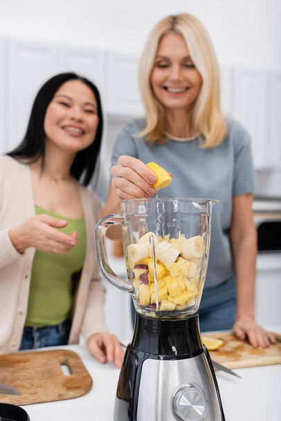 Zamazane międzyrasowe kobiety robiące koktajl owocowy w kuchni  - Zdjęcie, obraz