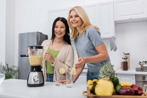 Cheerful interracial women looking away near smoothie and glasses in kitchen  - Fotoğraf, Görsel