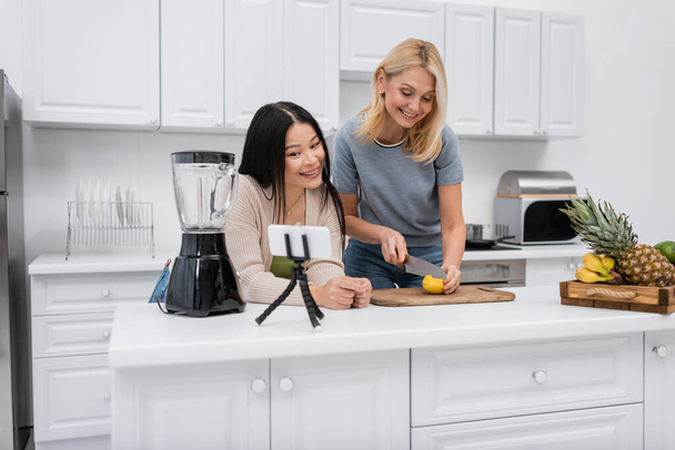 blogger multietnici che tagliano limone vicino smartphone su treppiede in cucina  - Foto, immagini