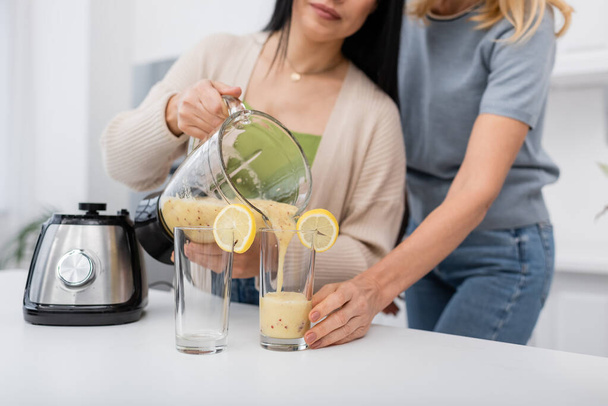 Gewassen uitzicht van de vrouw gieten smoothie in glas met citroen in de buurt wazig vriend thuis  - Foto, afbeelding