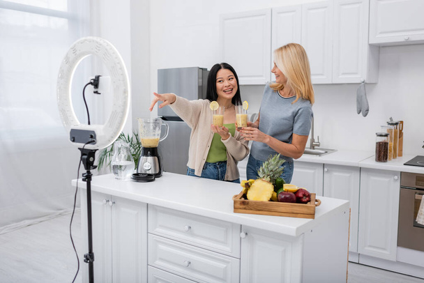Smiling asian blogger pointing at smartphone and ring light near friend with smoothie in kitchen  - Foto, imagen