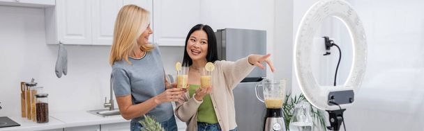 Cheerful asian blogger holding smoothie and pointing at smartphone in ring light near friend at home, banner  - Fotoğraf, Görsel