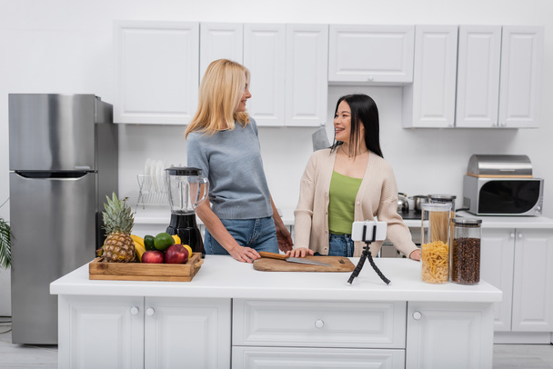 Positive interracial women standing near food and smartphone on tripod at home  - Photo, Image