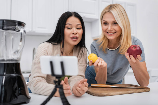 Cheerful woman holding fruits while asian friend talking at smartphone on tripod in kitchen  - Valokuva, kuva