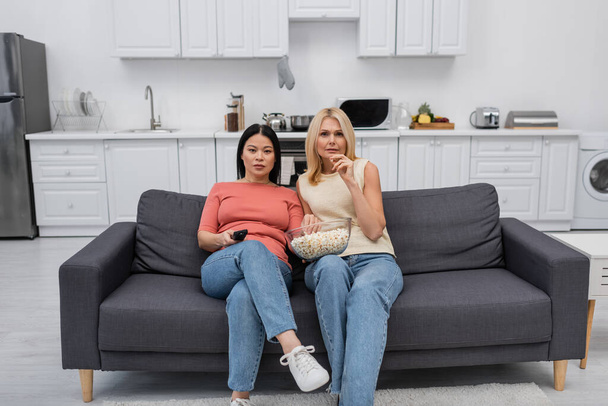 Interracial amigos segurando pipocas enquanto assiste tv no sofá em casa  - Foto, Imagem