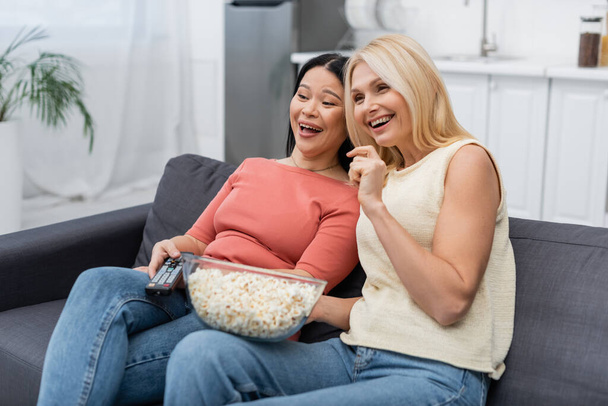 Fröhliche multiethnische Frauen mit Fernbedienung und Popcorn vor dem heimischen Fernseher  - Foto, Bild