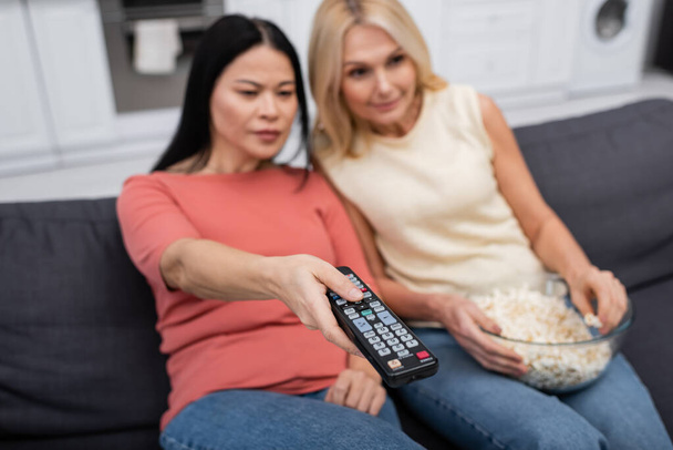 Blurred multiethnic women holding popcorn and clicking channels at home  - Photo, Image