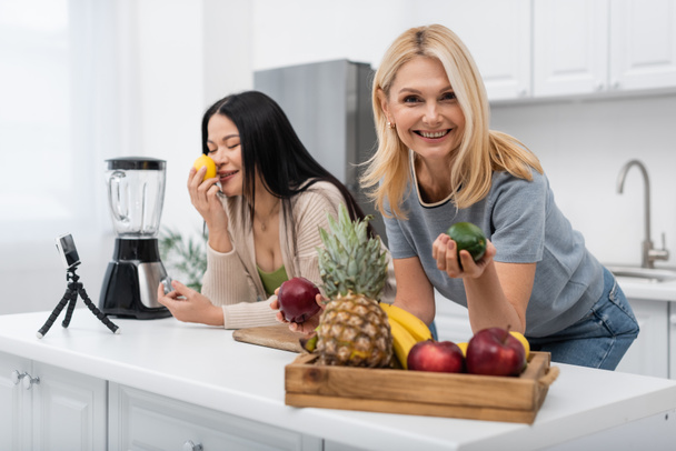 Sonriente mujer sosteniendo fruta cerca asiático amigo y smartphone en trípode en cocina  - Foto, imagen