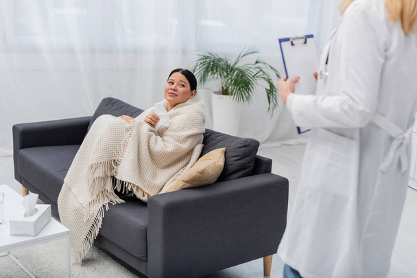 Sick asian woman holding napkin near blurred doctor with clipboard at home  - Foto, Bild