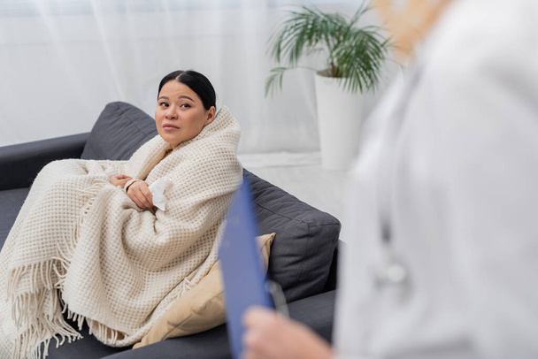 Sick asian patient in blanket looking at blurred doctor at home  - Foto, afbeelding