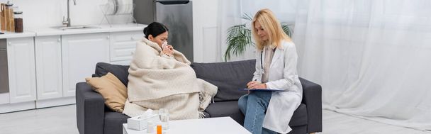 Doctor writing on clipboard near ill asian woman in blanket at home, banner  - Foto, afbeelding