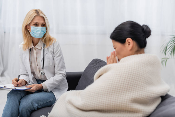 Doctor in medical mask holding clipboard near blurred asian patient at home  - Photo, Image