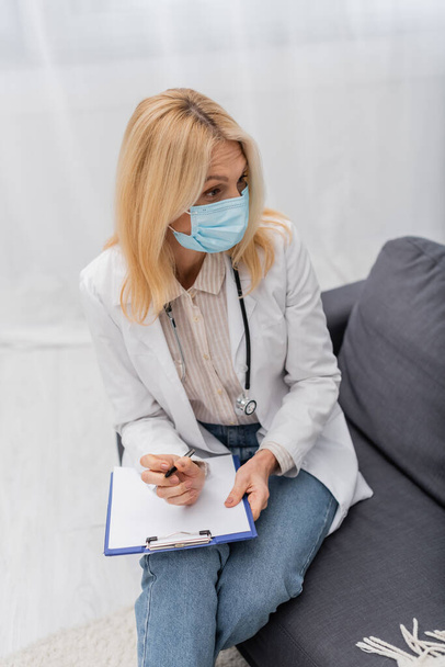 Mature doctor in medical mask holding clipboard and sitting on couch  - Photo, Image