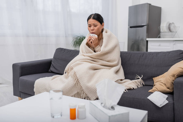 Уставшая и больная азиатка с салфеткой возле размытых таблеток дома  - Фото, изображение