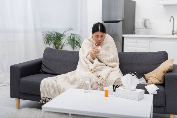 Verängstigte Asiatin in Decke schaut zu Hause auf Thermometer neben Tabletten und Servietten  - Foto, Bild