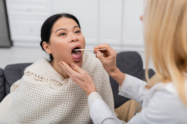 Blurred doctor en blanco capa chequeo garganta de asiático paciente en casa  - Foto, Imagen