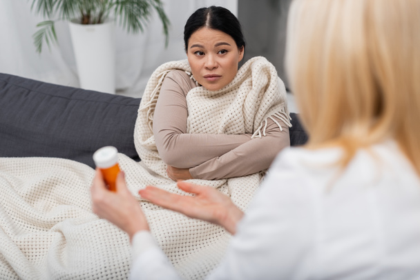 Zieke aziatische vrouw in deken kijken naar wazig arts wijzen naar pillen thuis  - Foto, afbeelding