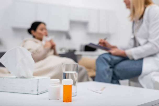 Таблетки и стакан воды возле размытого врача и пациента дома  - Фото, изображение