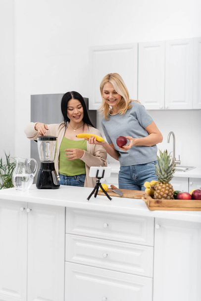 Positivi blogger interrazziali che tengono frutta vicino a frullatore e smartphone su treppiede a casa  - Foto, immagini
