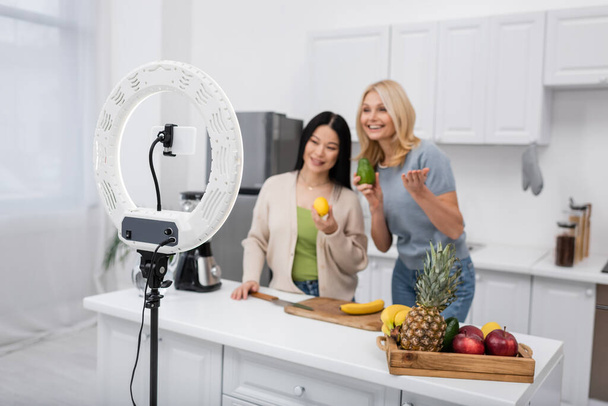 Кольцевая лампа и смартфон возле размытых межрасовых блогеров с спелыми фруктами дома  - Фото, изображение