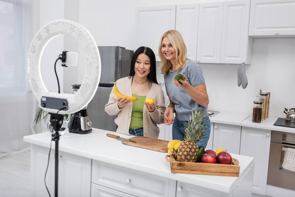 Multiethnic bloggers holding organic fruits near smartphone and ring lamp at home  - Photo, Image