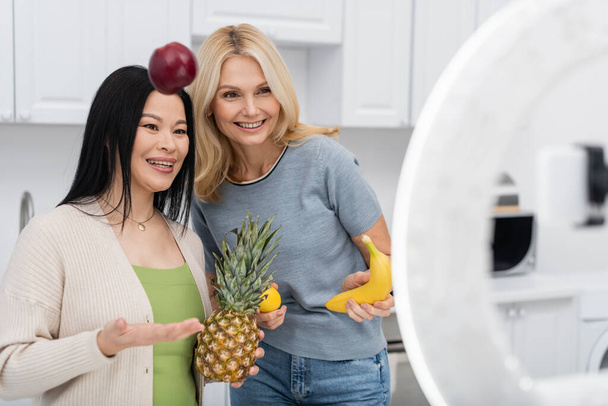 Blogger asiático lanzando manzana cerca de amigo y smartphone en cocina  - Foto, imagen